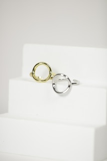 Two metal rings, one silver and one gold, are placed on a minimalist white staircase. The rings are modern and geometric in design, featuring round open shapes. The background is a plain, soft white, accentuating the elegance and simplicity of the objects.