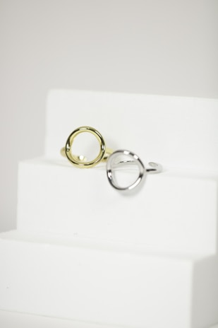 Two metal rings, one silver and one gold, are placed on a minimalist white staircase. The rings are modern and geometric in design, featuring round open shapes. The background is a plain, soft white, accentuating the elegance and simplicity of the objects.