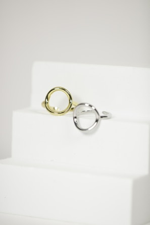 Two metal rings, one silver and one gold, are placed on a minimalist white staircase. The rings are modern and geometric in design, featuring round open shapes. The background is a plain, soft white, accentuating the elegance and simplicity of the objects.