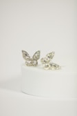 Two butterfly-shaped rings with intricate diamond patterns are elegantly displayed on a round, white pedestal. The rings have a delicate and shimmering appearance, with a focus on the sparkling gemstones that encrust each butterfly.