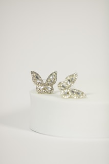 Two butterfly-shaped rings with intricate diamond patterns are elegantly displayed on a round, white pedestal. The rings have a delicate and shimmering appearance, with a focus on the sparkling gemstones that encrust each butterfly.