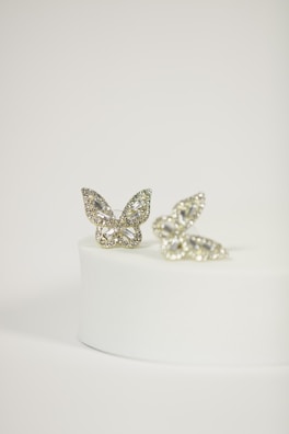Two butterfly-shaped rings with intricate diamond patterns are elegantly displayed on a round, white pedestal. The rings have a delicate and shimmering appearance, with a focus on the sparkling gemstones that encrust each butterfly.