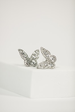 Two intricately designed butterfly-shaped earrings encrusted with small gemstones, placed on a white, minimalist platform. The earrings have a delicate and detailed appearance, with the light reflecting off their shiny surfaces.