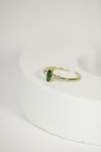 A delicate gold ring with a green gemstone at its center, flanked by two small diamonds, is placed on a rounded, light-colored surface. The focus is on the jewelry piece, showcasing its intricate design against a minimalistic background.