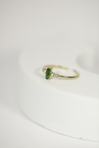 A delicate gold ring with a green gemstone at its center, flanked by two small diamonds, is placed on a rounded, light-colored surface. The focus is on the jewelry piece, showcasing its intricate design against a minimalistic background.