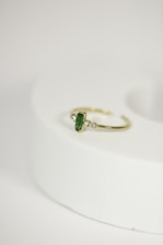A delicate gold ring with a green gemstone at its center, flanked by two small diamonds, is placed on a rounded, light-colored surface. The focus is on the jewelry piece, showcasing its intricate design against a minimalistic background.