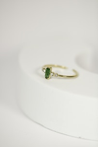 A delicate gold ring with a green gemstone at its center, flanked by two small diamonds, is placed on a rounded, light-colored surface. The focus is on the jewelry piece, showcasing its intricate design against a minimalistic background.