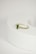 A delicate gold ring with a green gemstone at its center, flanked by two small diamonds, is placed on a rounded, light-colored surface. The focus is on the jewelry piece, showcasing its intricate design against a minimalistic background.