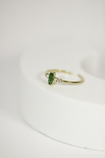 A delicate gold ring with a green gemstone at its center, flanked by two small diamonds, is placed on a rounded, light-colored surface. The focus is on the jewelry piece, showcasing its intricate design against a minimalistic background.