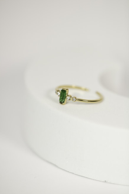 A delicate gold ring with a green gemstone at its center, flanked by two small diamonds, is placed on a rounded, light-colored surface. The focus is on the jewelry piece, showcasing its intricate design against a minimalistic background.