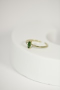 A delicate gold ring with a green gemstone at its center, flanked by two small diamonds, is placed on a rounded, light-colored surface. The focus is on the jewelry piece, showcasing its intricate design against a minimalistic background.