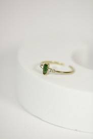 A delicate gold ring with a green gemstone at its center, flanked by two small diamonds, is placed on a rounded, light-colored surface. The focus is on the jewelry piece, showcasing its intricate design against a minimalistic background.
