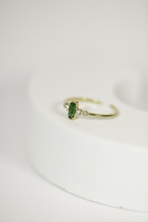 A delicate gold ring with a green gemstone at its center, flanked by two small diamonds, is placed on a rounded, light-colored surface. The focus is on the jewelry piece, showcasing its intricate design against a minimalistic background.
