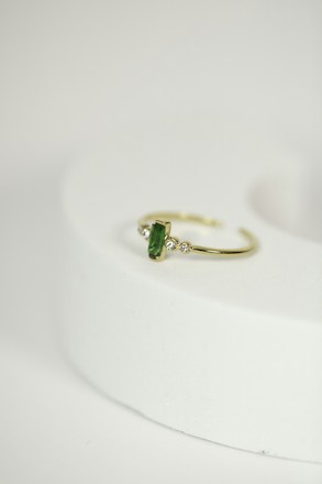A delicate gold ring with a green gemstone at its center, flanked by two small diamonds, is placed on a rounded, light-colored surface. The focus is on the jewelry piece, showcasing its intricate design against a minimalistic background.