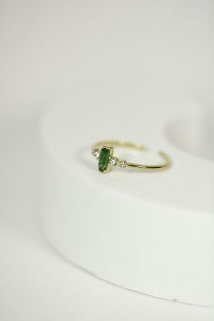 A delicate gold ring with a green gemstone at its center, flanked by two small diamonds, is placed on a rounded, light-colored surface. The focus is on the jewelry piece, showcasing its intricate design against a minimalistic background.