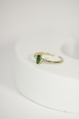 A delicate gold ring with a green gemstone at its center, flanked by two small diamonds, is placed on a rounded, light-colored surface. The focus is on the jewelry piece, showcasing its intricate design against a minimalistic background.