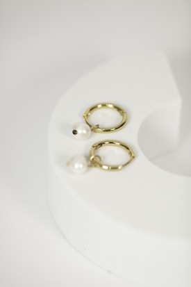 Two gold hoop earrings with white pearl pendants are placed on a white, curved, hollow object. The setup has a minimalist and elegant aesthetic.