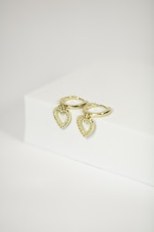 Two gold hoop earrings with heart-shaped pendants featuring small, sparkling stones are displayed on a white surface. The simple background highlights the elegance and shine of the jewelry.