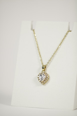 A gold necklace is adorned with a heart-shaped pendant featuring a central clear gemstone surrounded by smaller stones. The necklace is displayed on a plain white background, emphasizing its elegant design.