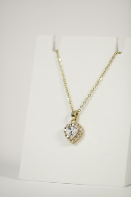 A gold necklace is adorned with a heart-shaped pendant featuring a central clear gemstone surrounded by smaller stones. The necklace is displayed on a plain white background, emphasizing its elegant design.