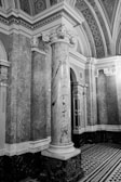 An intricately designed architectural interior featuring a large marble column with ornate carvings at its capital. The surrounding walls are covered with detailed stone marbling, and the ceiling is adorned with decorative patterns. The flooring features a geometric black-and-white tile design.