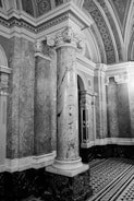 An intricately designed architectural interior featuring a large marble column with ornate carvings at its capital. The surrounding walls are covered with detailed stone marbling, and the ceiling is adorned with decorative patterns. The flooring features a geometric black-and-white tile design.