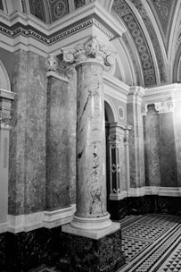 An intricately designed architectural interior featuring a large marble column with ornate carvings at its capital. The surrounding walls are covered with detailed stone marbling, and the ceiling is adorned with decorative patterns. The flooring features a geometric black-and-white tile design.