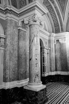 An intricately designed architectural interior featuring a large marble column with ornate carvings at its capital. The surrounding walls are covered with detailed stone marbling, and the ceiling is adorned with decorative patterns. The flooring features a geometric black-and-white tile design.