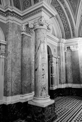 An intricately designed architectural interior featuring a large marble column with ornate carvings at its capital. The surrounding walls are covered with detailed stone marbling, and the ceiling is adorned with decorative patterns. The flooring features a geometric black-and-white tile design.