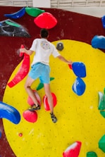 A person wearing a white shirt and blue shorts is climbing a colorful indoor bouldering wall. The wall features bright red, blue, yellow, and green climbing holds against a large yellow circular section on a dark red background. The person is gripping various holds with their hands and feet, showcasing physical activity and skill.