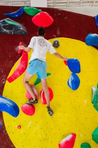 A person wearing a white shirt and blue shorts is climbing a colorful indoor bouldering wall. The wall features bright red, blue, yellow, and green climbing holds against a large yellow circular section on a dark red background. The person is gripping various holds with their hands and feet, showcasing physical activity and skill.