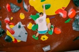 A person is bouldering indoors on an artificial climbing wall with various colorful holds. The wall has a mix of geometrically shaped holds in bright colors such as red, green, yellow, and black. The climber is wearing a white shirt with a colorful design and blue shorts, and is using both hands and feet to balance on the holds.