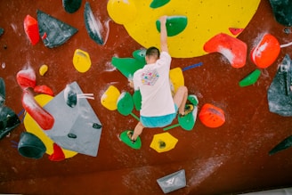 A person is bouldering indoors on an artificial climbing wall with various colorful holds. The wall has a mix of geometrically shaped holds in bright colors such as red, green, yellow, and black. The climber is wearing a white shirt with a colorful design and blue shorts, and is using both hands and feet to balance on the holds.
