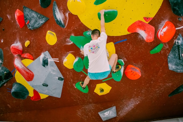 A person is bouldering indoors on an artificial climbing wall with various colorful holds. The wall has a mix of geometrically shaped holds in bright colors such as red, green, yellow, and black. The climber is wearing a white shirt with a colorful design and blue shorts, and is using both hands and feet to balance on the holds.