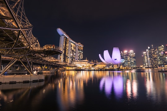 The image captures a stunning nighttime cityscape with modern architectural landmarks. A prominent curved structure resembling a futuristic bridge is visible on the left, while the center showcases an iconic building with a unique, illuminated lotus flower design. Skyscrapers with bright lights reflecting on the tranquil water create a vibrant and bustling urban feel.