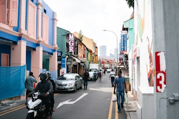 A vibrant city street scene featuring colorful buildings on both sides, with a mix of shops and stores visible. People walk along the sidewalk while a motorcyclist and a car travel down the street. Street signs and advertisements create a bustling, lively atmosphere.