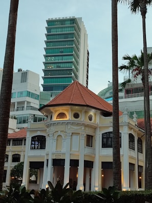 A colonial-style building with a prominent red-tiled roof stands in front of modern high-rise structures. Tall palm trees frame the scene, creating a contrast between the historical and the contemporary.
