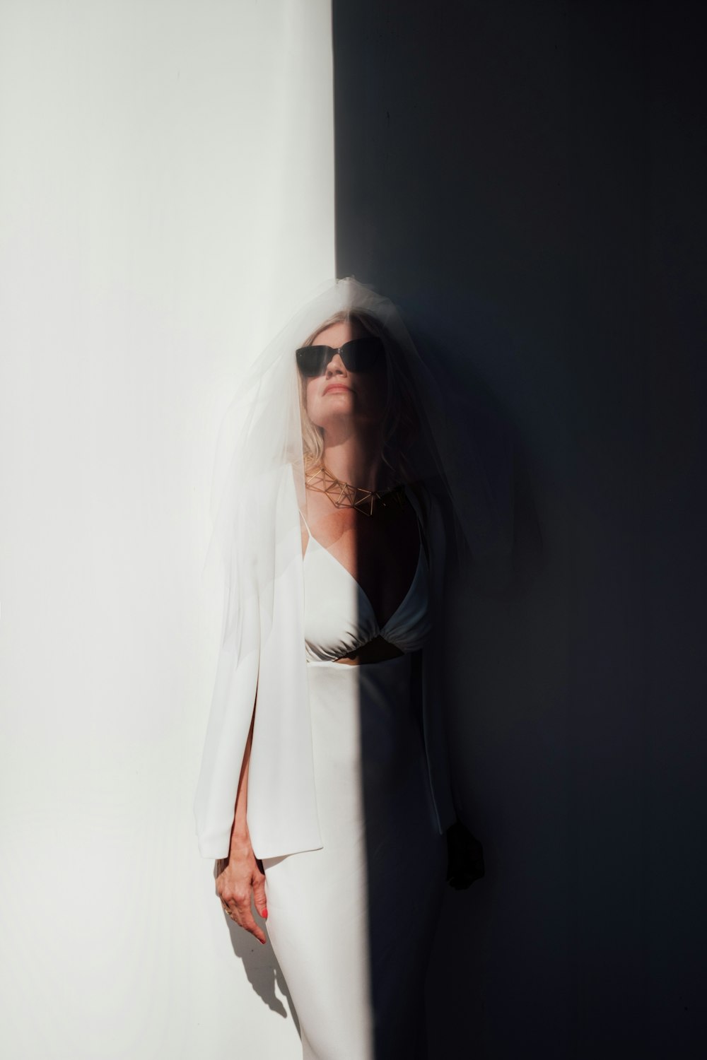 a woman in a white suit and sunglasses leaning against a wall