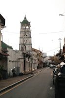 A narrow street lined with weathered buildings and a tall, historic tower featuring multiple levels with arches and a green roof at the top. There are power lines crossing overhead, a parked motorcycle, and a few cars along the street. The architecture reflects a colonial style, and a bird is flying in the sky.