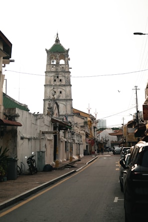 A narrow street lined with weathered buildings and a tall, historic tower featuring multiple levels with arches and a green roof at the top. There are power lines crossing overhead, a parked motorcycle, and a few cars along the street. The architecture reflects a colonial style, and a bird is flying in the sky.