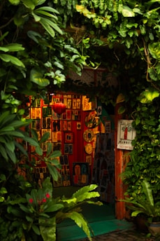 A cozy, vibrant art gallery entrance adorned with lush green foliage. Various colorful artworks are displayed on the walls, featuring abstract and figurative designs. Bright red, orange, and yellow hues create a warm ambiance, while a few small clocks and a red lantern add character to the setting.