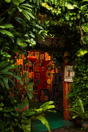 A cozy, vibrant art gallery entrance adorned with lush green foliage. Various colorful artworks are displayed on the walls, featuring abstract and figurative designs. Bright red, orange, and yellow hues create a warm ambiance, while a few small clocks and a red lantern add character to the setting.