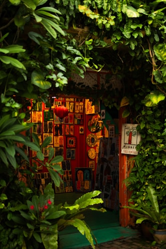 A cozy, vibrant art gallery entrance adorned with lush green foliage. Various colorful artworks are displayed on the walls, featuring abstract and figurative designs. Bright red, orange, and yellow hues create a warm ambiance, while a few small clocks and a red lantern add character to the setting.