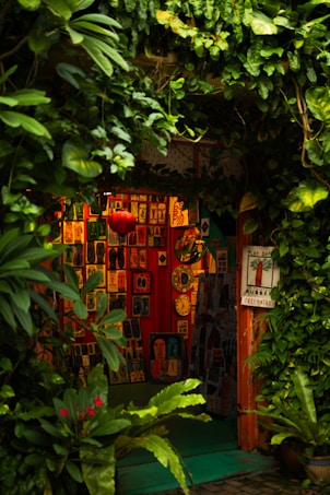 A cozy, vibrant art gallery entrance adorned with lush green foliage. Various colorful artworks are displayed on the walls, featuring abstract and figurative designs. Bright red, orange, and yellow hues create a warm ambiance, while a few small clocks and a red lantern add character to the setting.