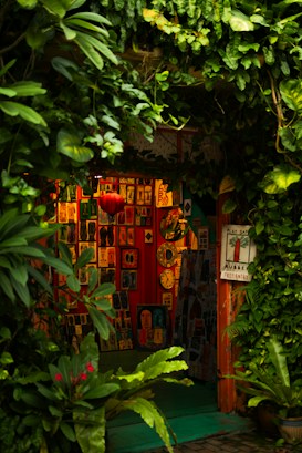A cozy, vibrant art gallery entrance adorned with lush green foliage. Various colorful artworks are displayed on the walls, featuring abstract and figurative designs. Bright red, orange, and yellow hues create a warm ambiance, while a few small clocks and a red lantern add character to the setting.