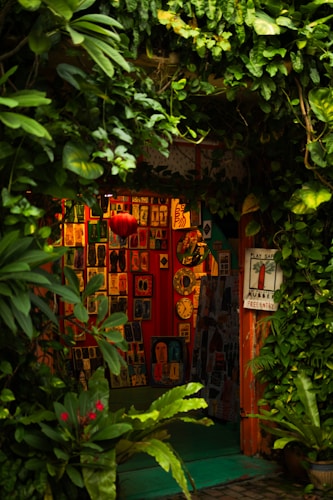 A cozy, vibrant art gallery entrance adorned with lush green foliage. Various colorful artworks are displayed on the walls, featuring abstract and figurative designs. Bright red, orange, and yellow hues create a warm ambiance, while a few small clocks and a red lantern add character to the setting.