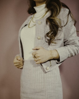 A woman in a tailored, pastel-colored suit jacket and skirt, accessorized with gold jewelry, including a necklace and a bracelet. Her long, wavy brown hair falls over her shoulder, and a soft pink background complements the outfit.