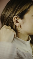 A close-up of a person wearing delicate gold jewelry, including ear cuffs and hoop earrings. The individual has their hand near their neck, wearing a ring, and is dressed in a soft, light-colored sweater.