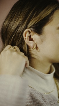 A close-up of a person wearing delicate gold jewelry, including ear cuffs and hoop earrings. The individual has their hand near their neck, wearing a ring, and is dressed in a soft, light-colored sweater.