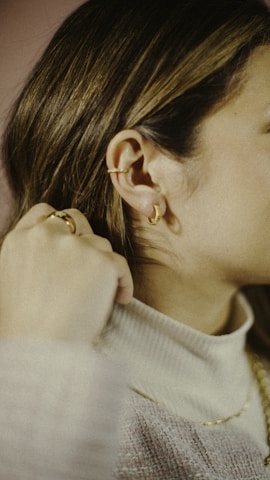 A close-up of a person wearing delicate gold jewelry, including ear cuffs and hoop earrings. The individual has their hand near their neck, wearing a ring, and is dressed in a soft, light-colored sweater.